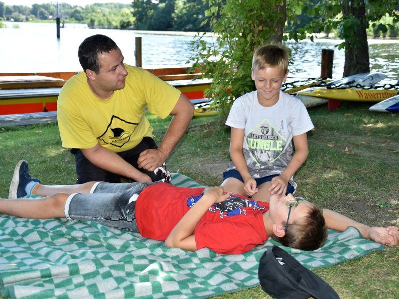 Ein Kind liegt auf einer Decke in der Wiese. Ein zweites Kind kniet daneben und sie üben gemeinsam die stabile Seitenlage. Dabei werden Sie durch einen Betreuer beobachtet und angeleitet.