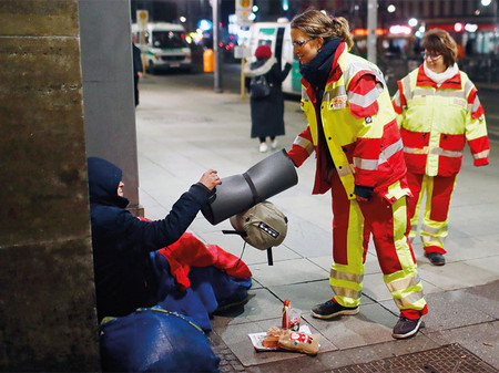 Samariterin überreicht Wohnungslosem Schlafsack und Isomatte.